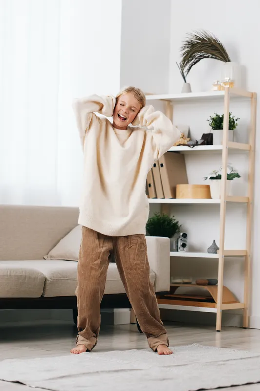 Young person stretching cheerfully in a bright living room, wearing a soft cream sweater and cozy patterned lounge pants, capturing a casual, comfortable home moment of daily life.