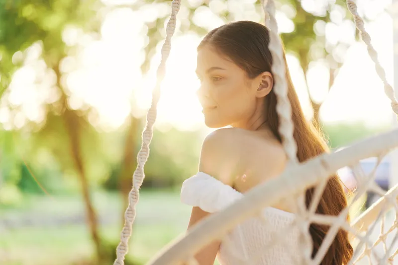 A serene young woman enjoying a moment of relaxation in a swing, showcasing natural beauty and tranquility in a sunny outdoor setting