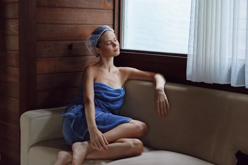 woman relaxing wrapped in a blue towel sitting on a beige sofa near a window with sheer curtains in soft natural light