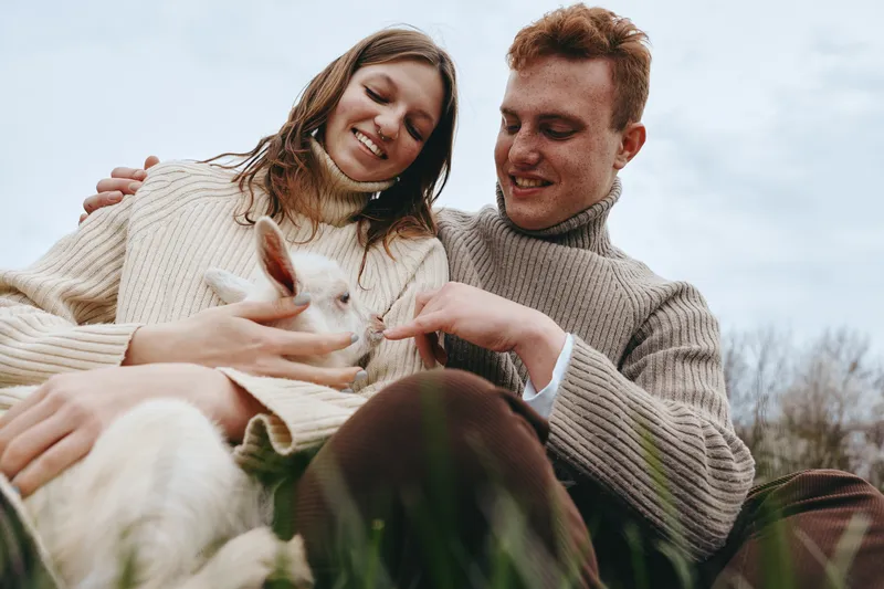 Smiling couple outdoors with a goat kid, cozy sweaters, relaxed mood, close interaction, nature setting, candid lifestyle moment, warm tones, affection and companionship, simple happiness, rustic