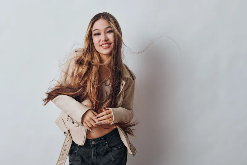 Young woman with long hair wearing beige jacket and black pants smiling and posing against plain background. Casual fashion, portrait, beauty, and confident expression with natural light.