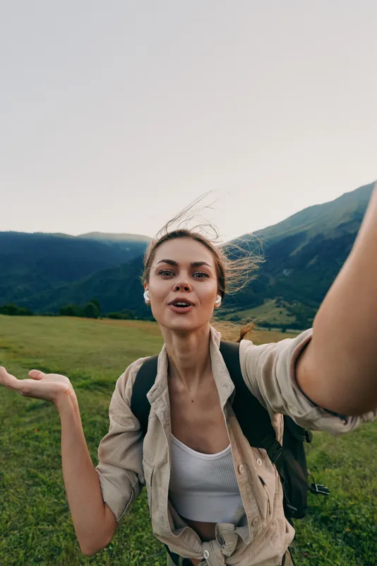 Young woman with a backpack takes a selfie while hiking in the mountains on a clear day with green grass and blue hills in the background.