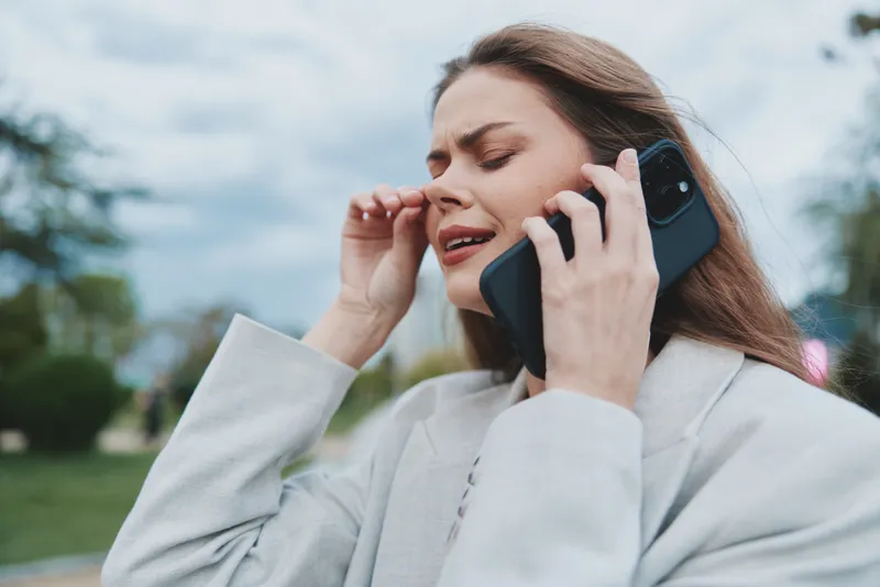 Businesswoman distressed while on the phone, wearing a light gray suit, outdoors with an overcast sky, conveying stress and urgency amidst a professional backdrop.