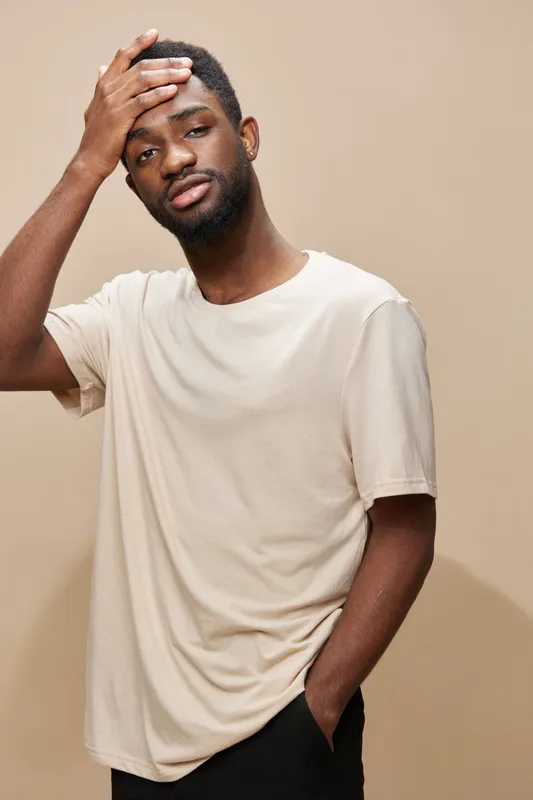 Young Black man in beige T shirt expressing contemplation against a neutral background, reflecting emotions of thoughtfulness and introspection