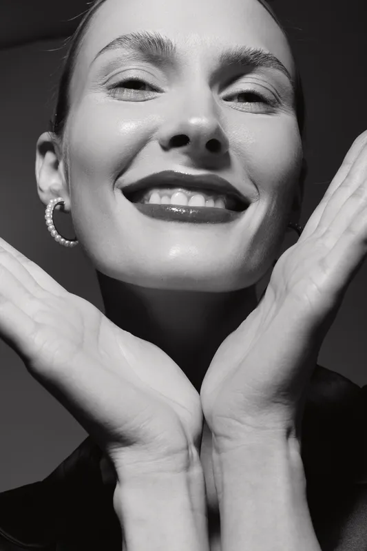 Close up of smiling white woman with light skin and earrings posing with hands near face. Studio portrait in black and white with soft lighting and joyful expression.