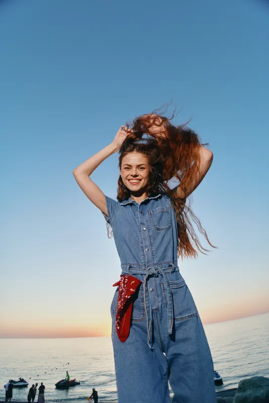 lifestyle woman in modern boho-western denim street style enjoying sunset by sea, smiling with flowing hair in warm film color tones, casual and natural vibe