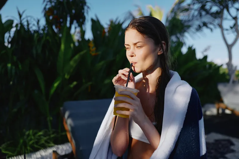 Cheerful woman outdoors, relaxing with a drink, sunlit moment amid lush greenery, gentle breeze on skin, towel draped over shoulders, casual summer lifestyle, smiling softly, enjoying fresh air and
