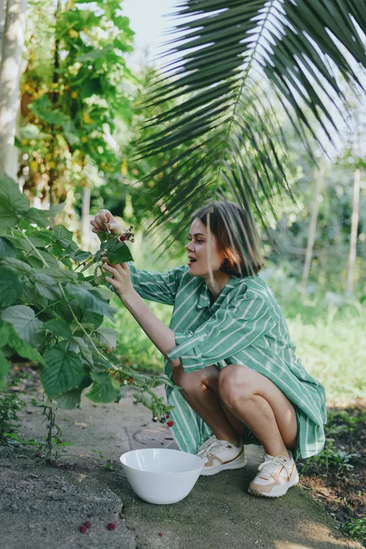 Young woman crouches in a lush garden, picking ripe berries into a white bowl, surrounded by tropical leaves and warm sunlight.