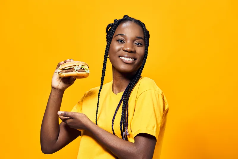 Young Black woman in a yellow shirt smiling joyfully while holding a delicious sandwich against a vibrant yellow background, highlighting healthy eating trends