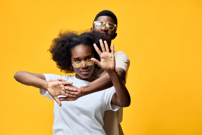 Young African man and woman posing together against a bright yellow background, showcasing playful emotions and modern fashion style