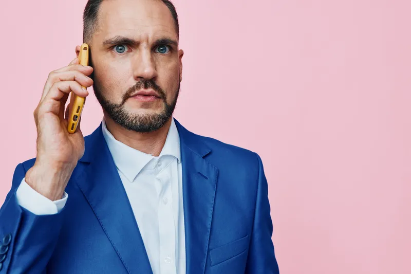 A focused business professional speaks on a mobile phone against a solid pink background, conveying concentration, urgency, and determination in a modern corporate mood.