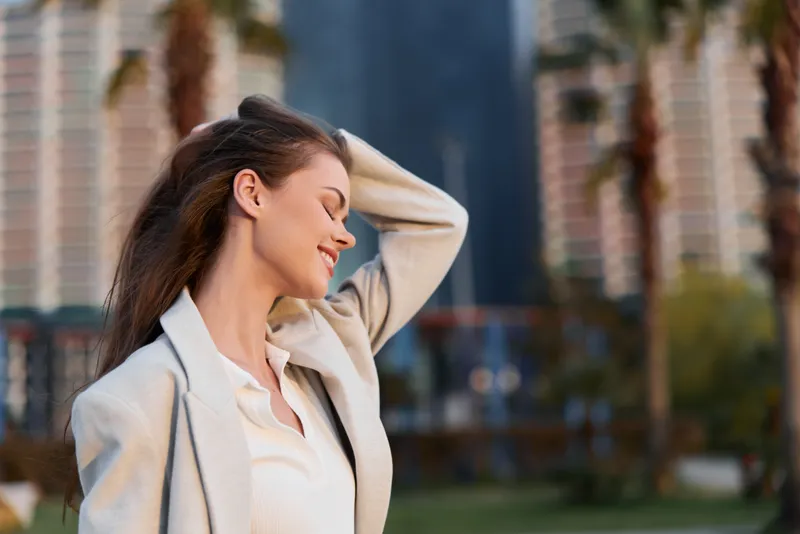 Confident woman in business attire smiling outdoors, captured in a vibrant urban setting with palm trees, showcasing a professional yet relaxed atmosphere.