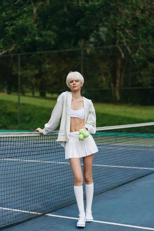 Young female tennis player in a sporty outfit, exuding confidence with a smile, stands by the net on an outdoor court surrounded by lush greenery