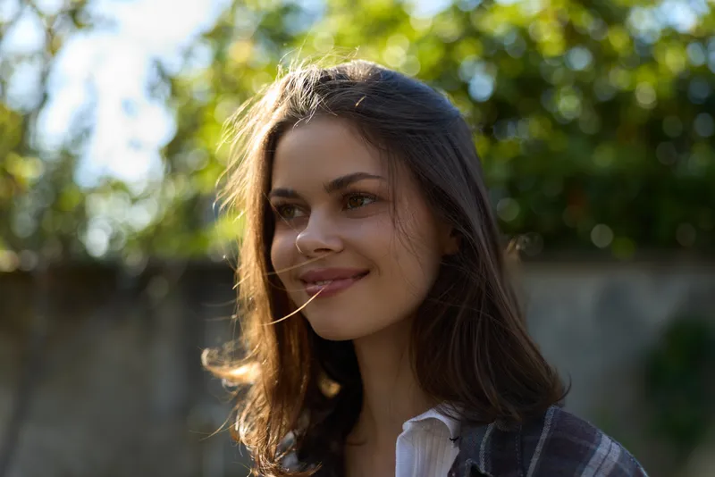 A young woman with long brunette hair smiles softly outdoors in warm sunlight, standing near a leafy fence with a relaxed, natural pose and gentle breeze