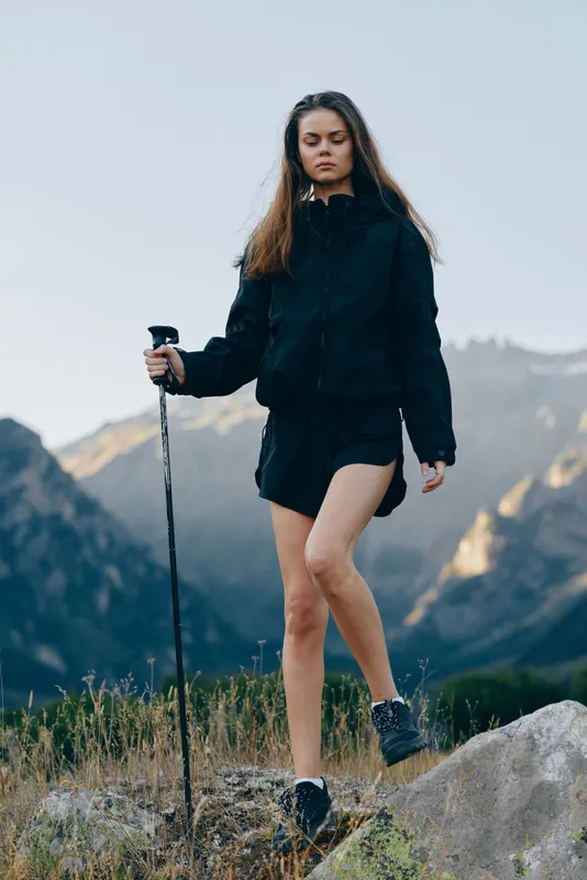 A focused hiker woman navigates a rocky trail in a rugged mountain landscape. She wears a dark jacket and shorts, using a trekking pole, exuding adventure and determination.