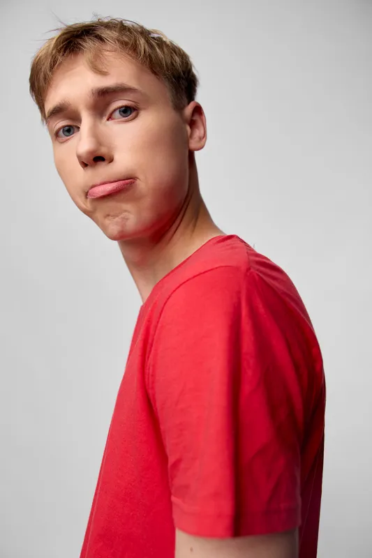 Young man in a red t shirt with a puzzled expression, half turned against a neutral studio background, conveying confusion, casual fashion and moody attitude.