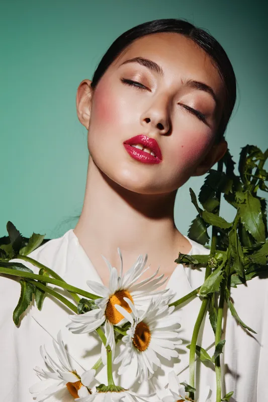 Young woman with glossy red lips and closed eyes poses with white daisies and green leaves on her shoulders against a soft green background. Beauty, fashion, artistic concept.