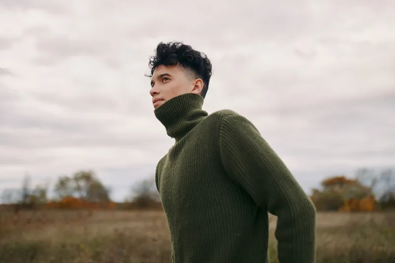 Young man in a green turtleneck sweater stands in a windswept field, captured with authenticity and realism using natural light to emphasize genuine presence and quiet confidence.