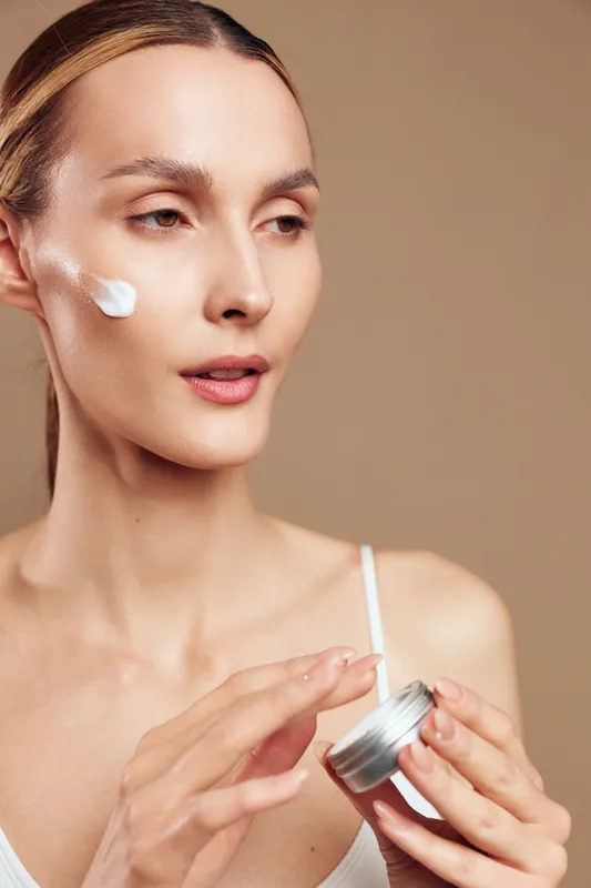 Young woman applies facial cream on cheek with fingers holding jar skincare beauty routine natural fresh healthy skin closeup portrait on beige background.