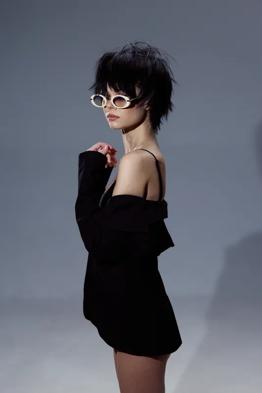 Stylish woman wearing white sunglasses and black outfit posing against gray background with moody lighting and modern haircut.