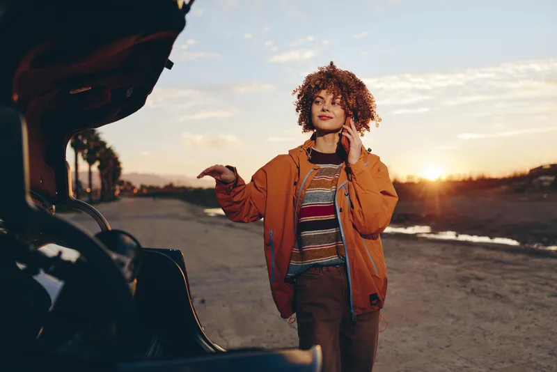Woman lifestyle outdoors at sunset, curly hair woman by open car door, hand raised while talking, warm evening light, casual jacket and jeans, travel mood on road after work soft breeze