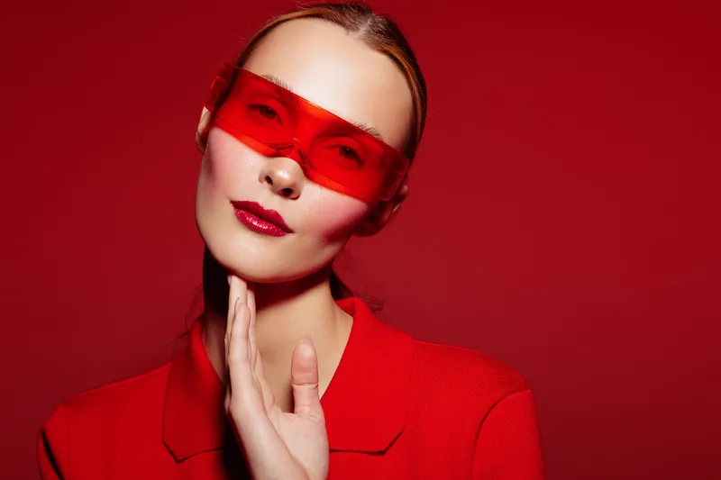 Stylish woman in red coat and red translucent glasses poses against red background. Fashion portrait with bold makeup and confident expression. Concept of modern style and elegance.