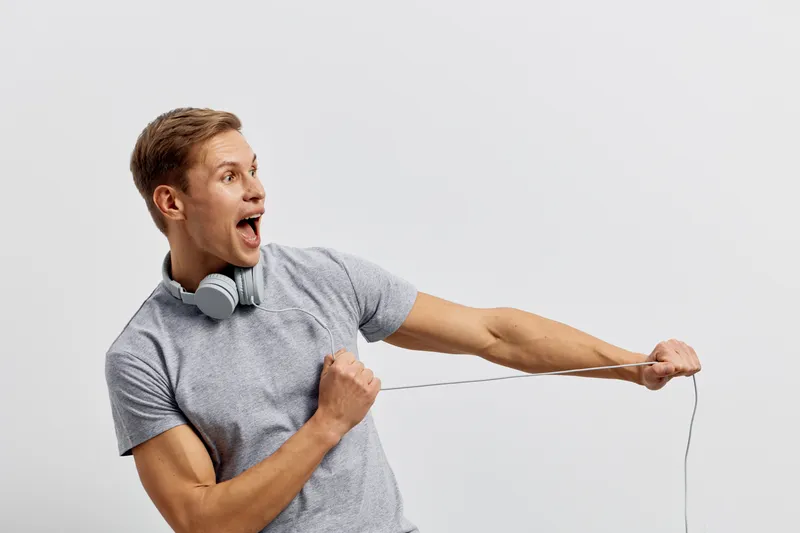 Energetic young man with headphones listens to music, enthusiastically singing and pulling a headphone wire, excited expression, casual grey t-shirt, standing against a plain light background, lifesty