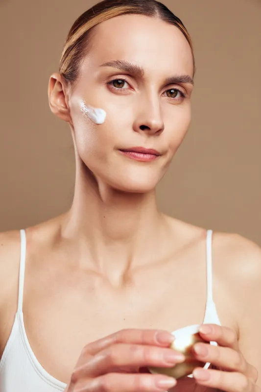 Young woman applies facial cream on clean skin holds jar looks at camera with calm expression isolated on brown background skincare beauty routine concept natural care