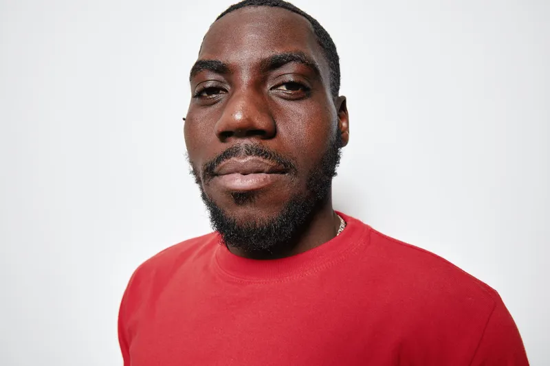Portrait of African American man with dark skin wearing red shirt looking confidently at camera on white background. Closeup, facial hair and casual style with natural expression.