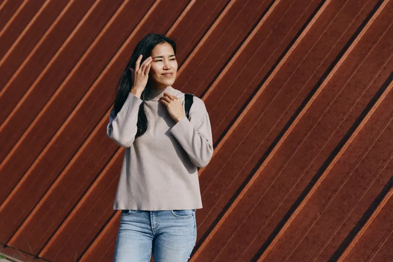 Woman with backpack talking on smartphone outdoors against a diagonal wooden wall, casual sweater and jeans, lifestyle moment showing urban style, happy mood and everyday communication in a travel