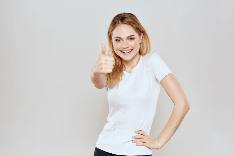 Весёлая блондинка в белой футболке жестикулирует руками на светлом фоне