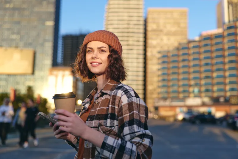 Woman holding coffee and phone in urban street at golden hour sunlight, smile framed by beanie and casual outfit. Candid lifestyle portrait emphasizing authenticity, mindful living and emotional