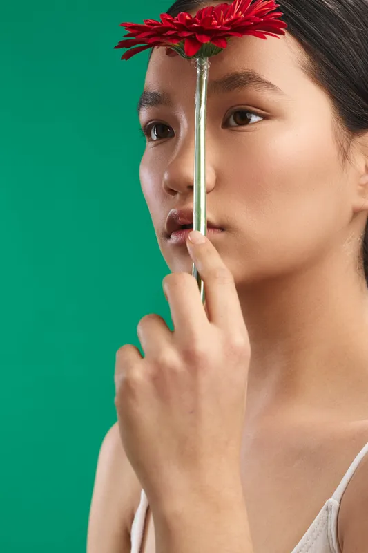 Asian woman is photographed in a close up portrait holding a red flower stem across her face. Soft yet bold lighting enhances calm, mood, and artistic expression.