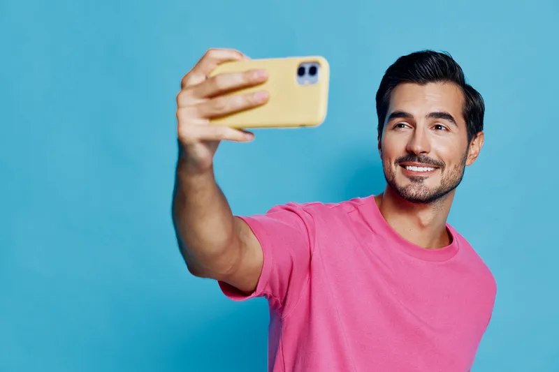 Smiling man taking selfie with mobile phone, wearing pink t-shirt against bright blue background, enjoying moment of happiness and self-expression in a cheerful atmosphere.