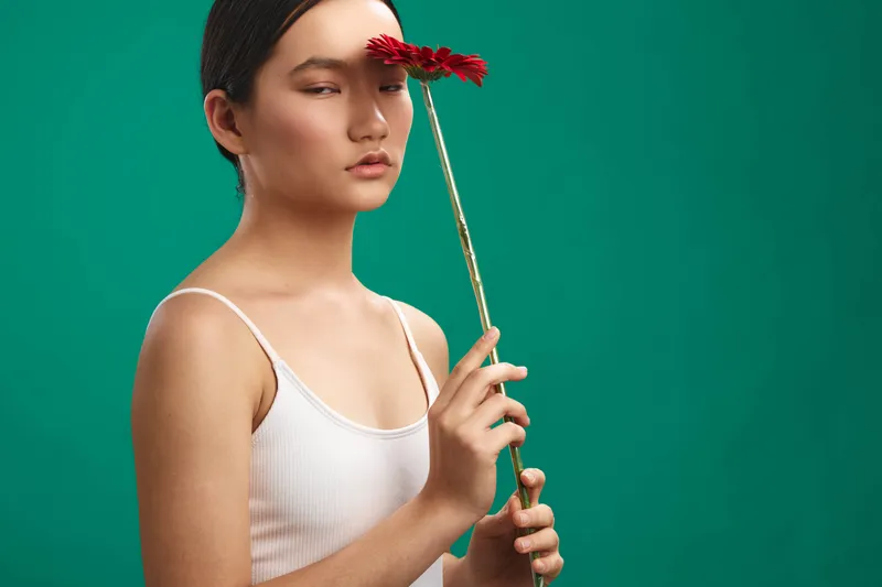 asian woman holds a flower against a teal backdrop in a minimalist studio. she wears a white tank, gazes softly, and exudes calm, focus, and subtle grace in the portrait.