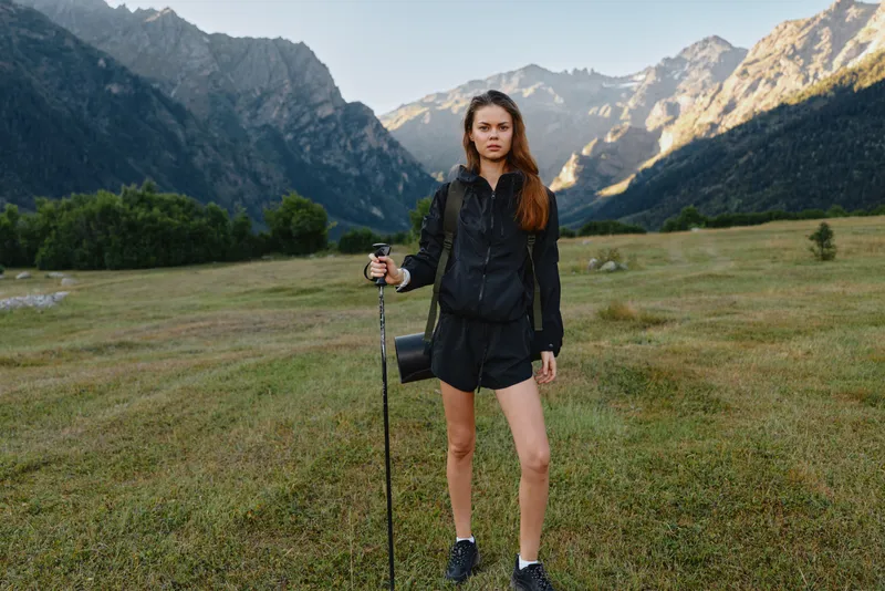 A confident young hiker stands in a grassy valley with a walking staff, dressed in black outdoor gear, framed by dramatic mountains under a clear sky.
