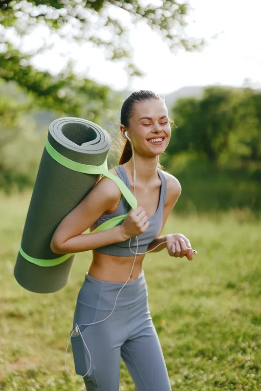 Happy young woman in activewear carrying yoga mat in nature, embodying a healthy lifestyle and fitness concept
