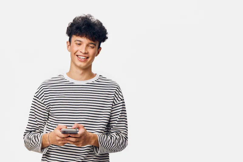 smiling man in striped shirt holding smartphone, standing against white background with curly hair, casual and modern appearance, youthful and friendly expression