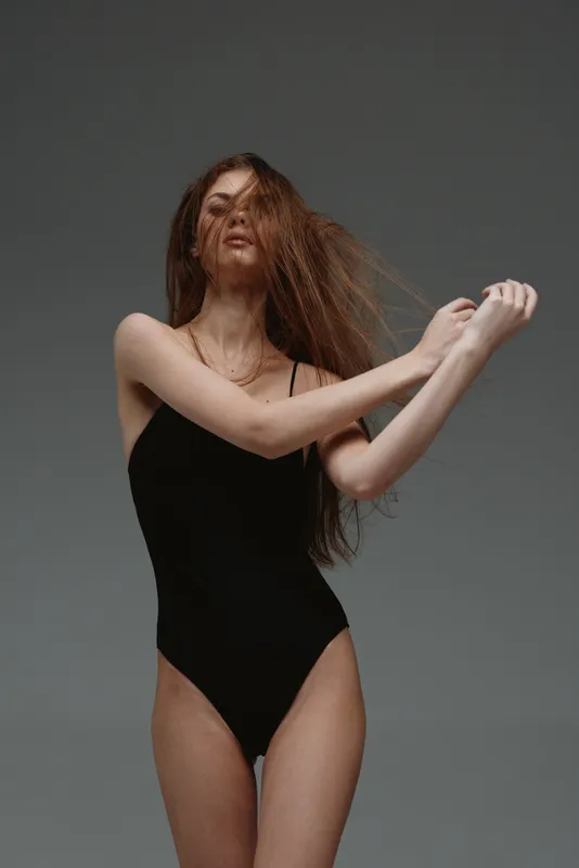 slender woman wearing black bodysuit posing with long hair in studio against gray background