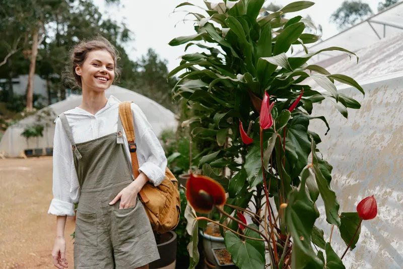 Young woman smiling in trendy overalls, walking among tropical plants in a greenhouse, showcasing joy and nature s beauty in vibrant colors