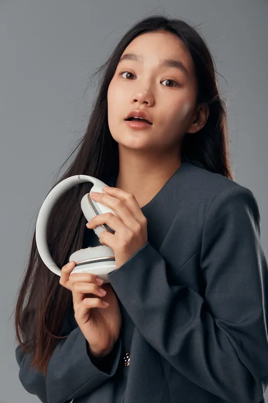 Young woman with long dark hair holds white headphones in hands looking forward with open mouth, wearing dark gray jacket on gray background in studio portrait.