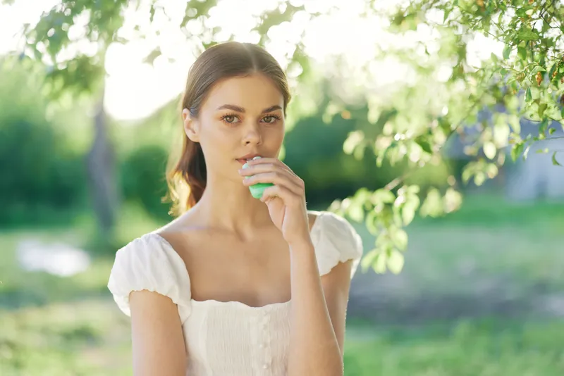 Young woman in a white dress holding a green smoothie, surrounded by greenery, conveying freshness and healthy lifestyle in a seasonal outdoor setting