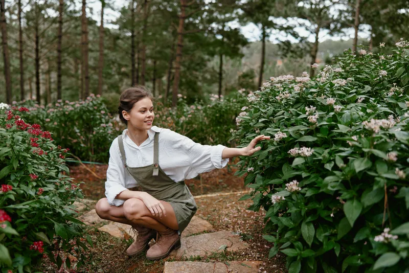 Young woman in overalls enjoying nature in a garden, surrounded by blooming flowers and greenery, evoking feelings of tranquility, joy, and connection with nature