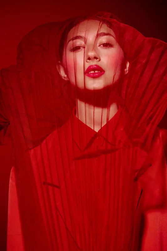 Portrait of a young woman dressed in red fashion attire with dramatic veil over her face. Bold red lipstick enhances the creative beauty concept with artistic lighting effects.