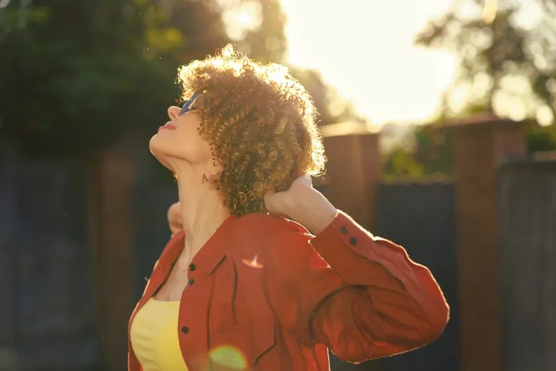 A woman with curly hair in a red jacket gazes upward in bright sunlight, conveying outdoor lifestyle, happiness, and a contemplative moment in a warm, relaxed setting, showcasing wellbeing and self