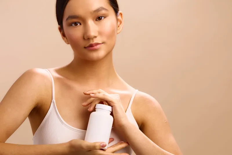 asian woman with a calm expression holds a white skincare bottle in a soft studio setting, presenting a clean, modern vibe and a focus on self care, wellness, and gentle daily beauty ritual.