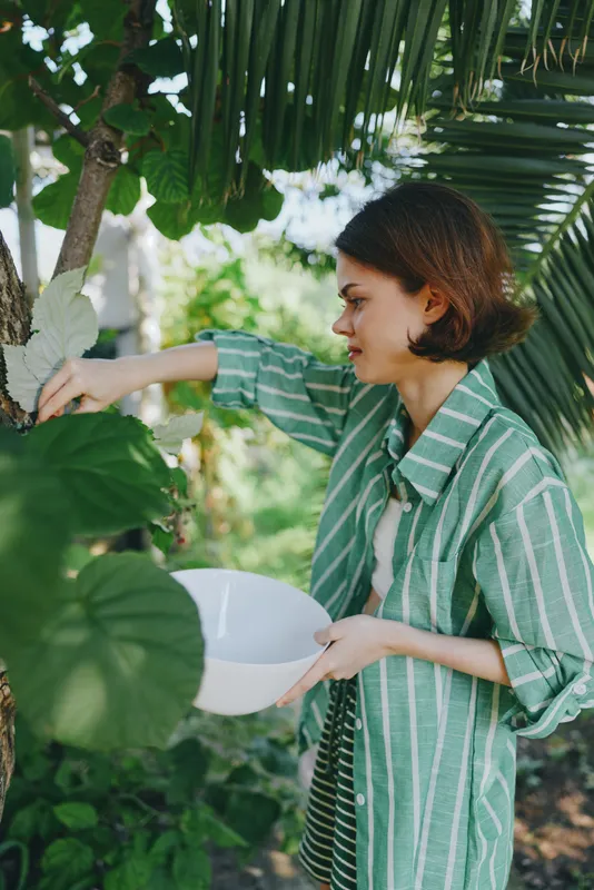 Young woman tends tropical plants, reaching to inspect leaves while holding a white bowl, surrounded by lush greenery.