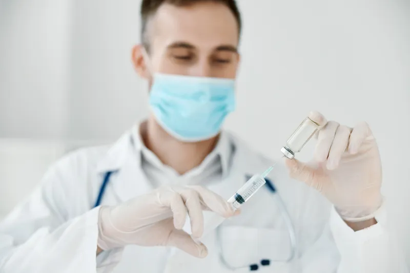 Врач в медицинской маске держит в руке шприц с вакциной, химической жидкостью, рядом стетоскоп