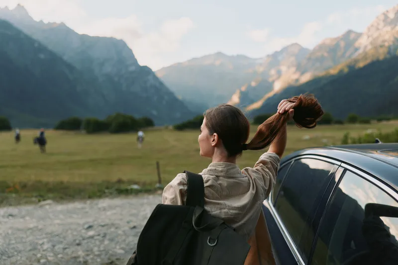 A woman lifestyle traveler stands next to a car in a mountain valley holding her hair up while looking at the distant peaks under a cloudy sky.