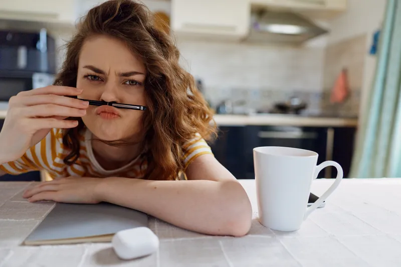 Young woman in a striped shirt sits at a kitchen table, playfully holding a pen under her nose, looking tired yet curious and slightly amused.